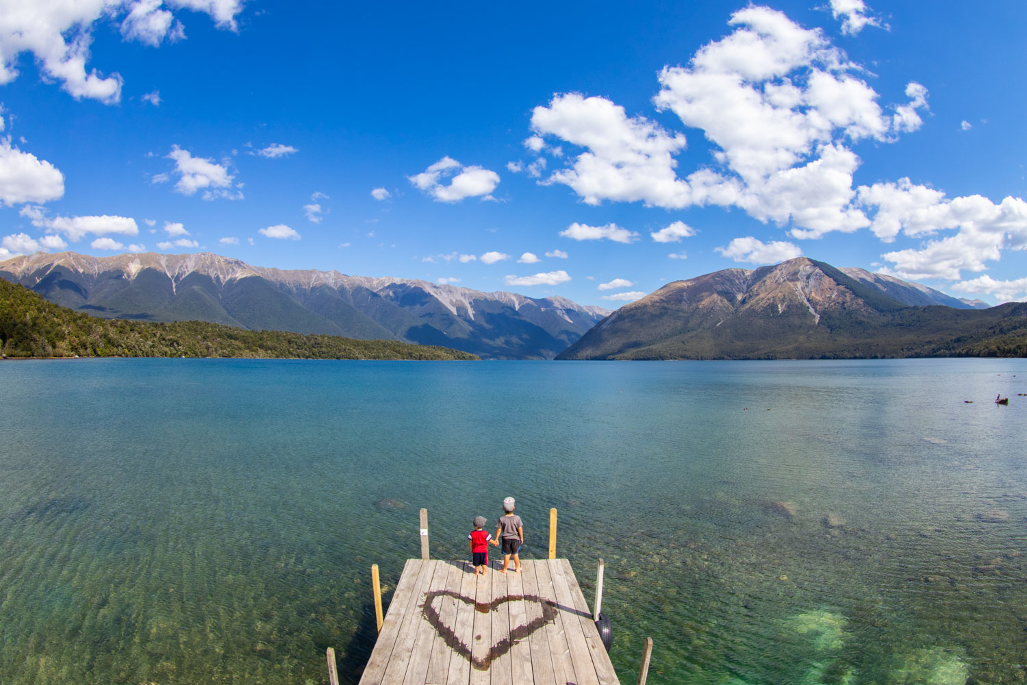 Debout sur un quai en bois, deux enfants se tiennent la main au bord d’un lac et regardent les montagnes au loin.