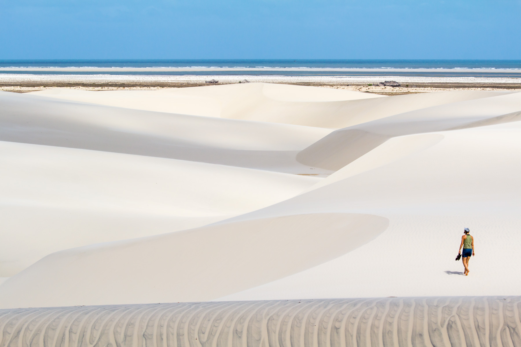 Désert et dunes de sable où une femme marche pied nu de dos. À l’horizon, on aperçoit l’océan.