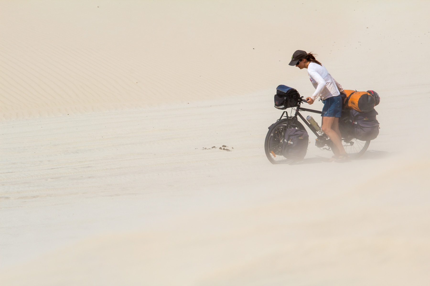 Près d’une dune, une femme dans la trentaine pousse un vélo chargé dans le sable.