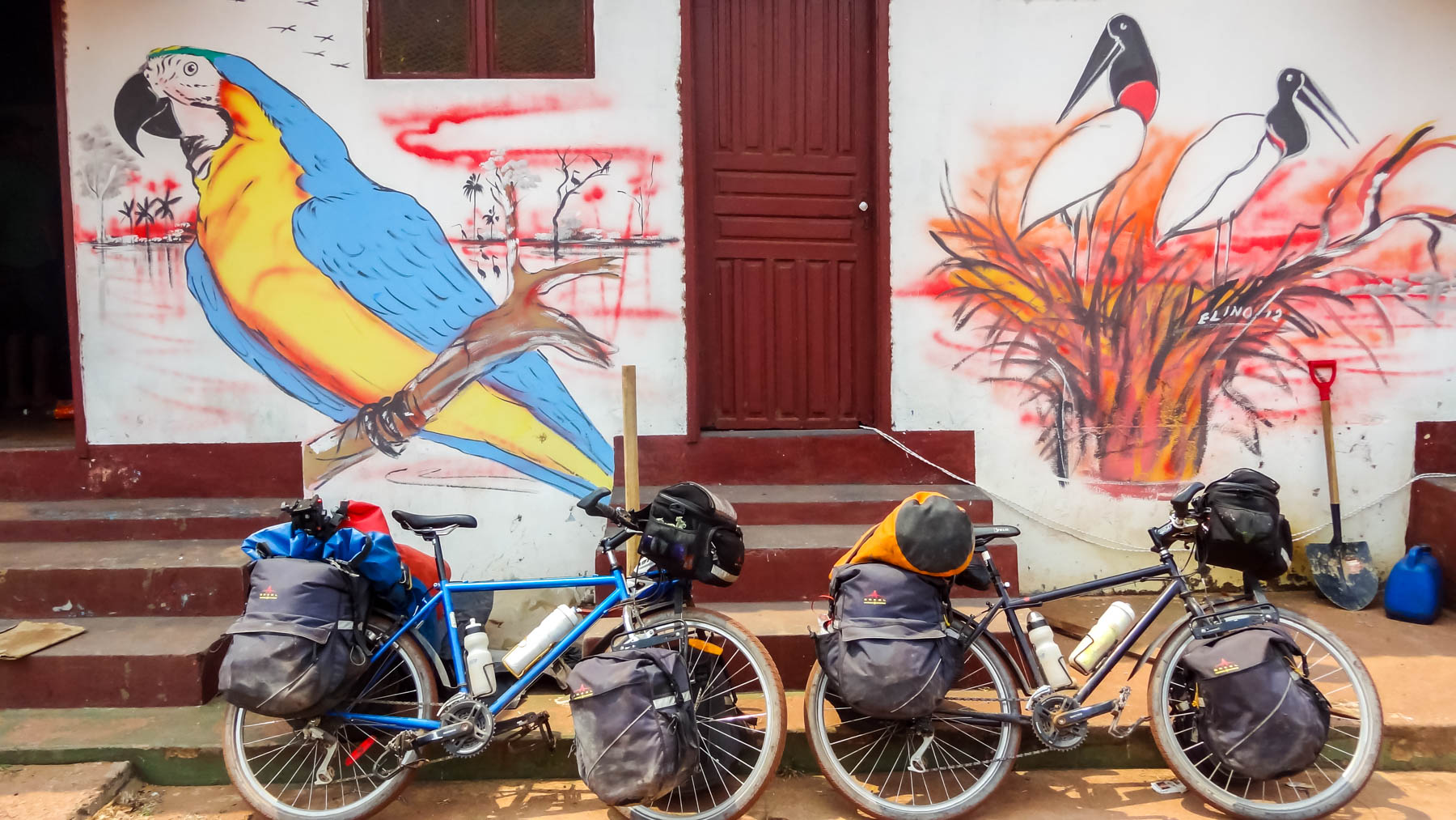 Deux vélos de cyclotourisme accotés près d’une maison ornée de grandes fresques d’oiseaux exotiques.
