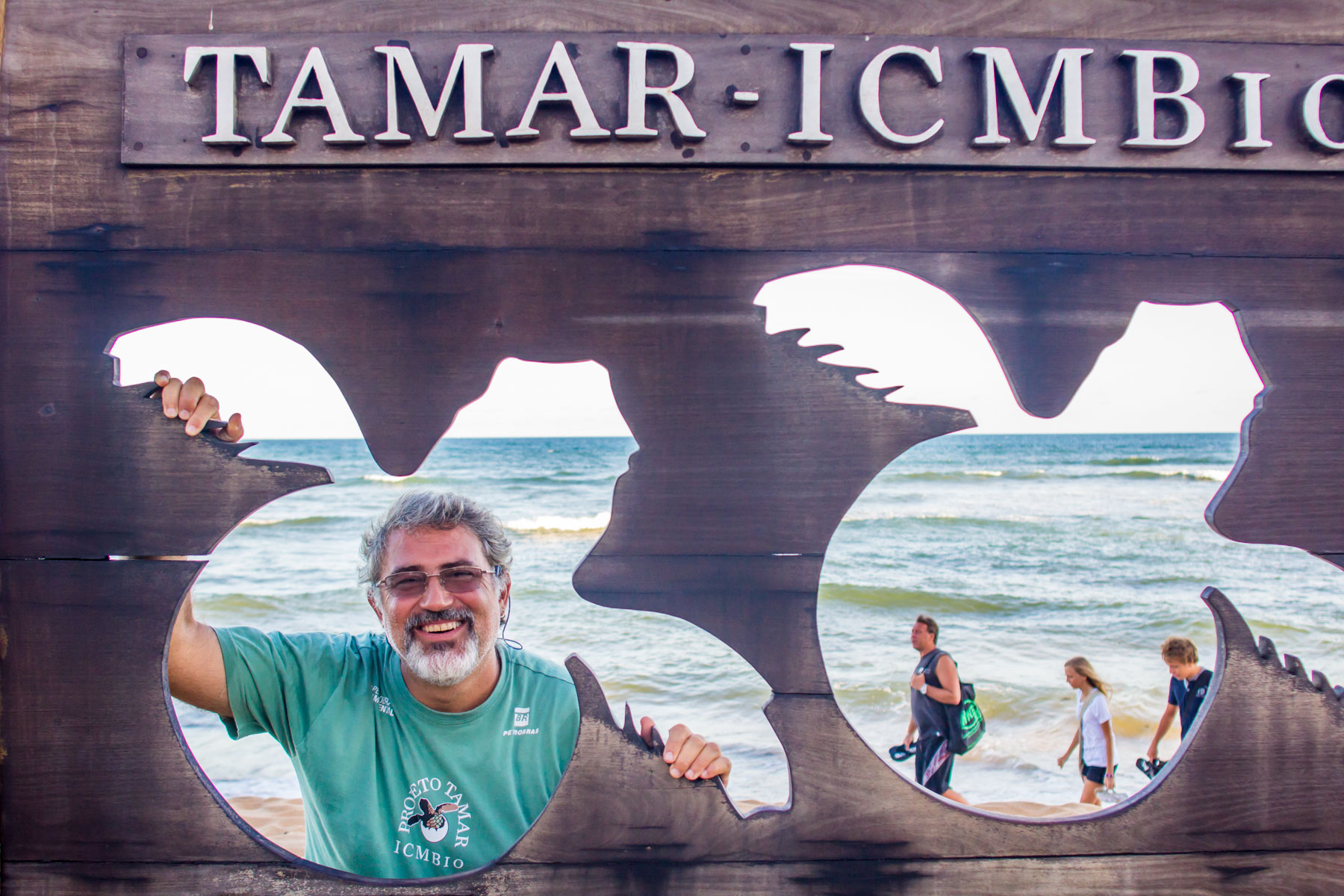 At the ocean’s edge and behind a wooden sign in the shape of a turtle, a man with a beard and white hair smiles at the photographer.