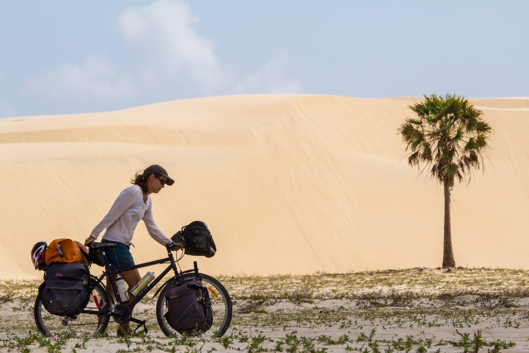 Près d’une dune et d’un palmier, une femme dans la trentaine pousse un vélo chargé dans un chemin sablonneux.