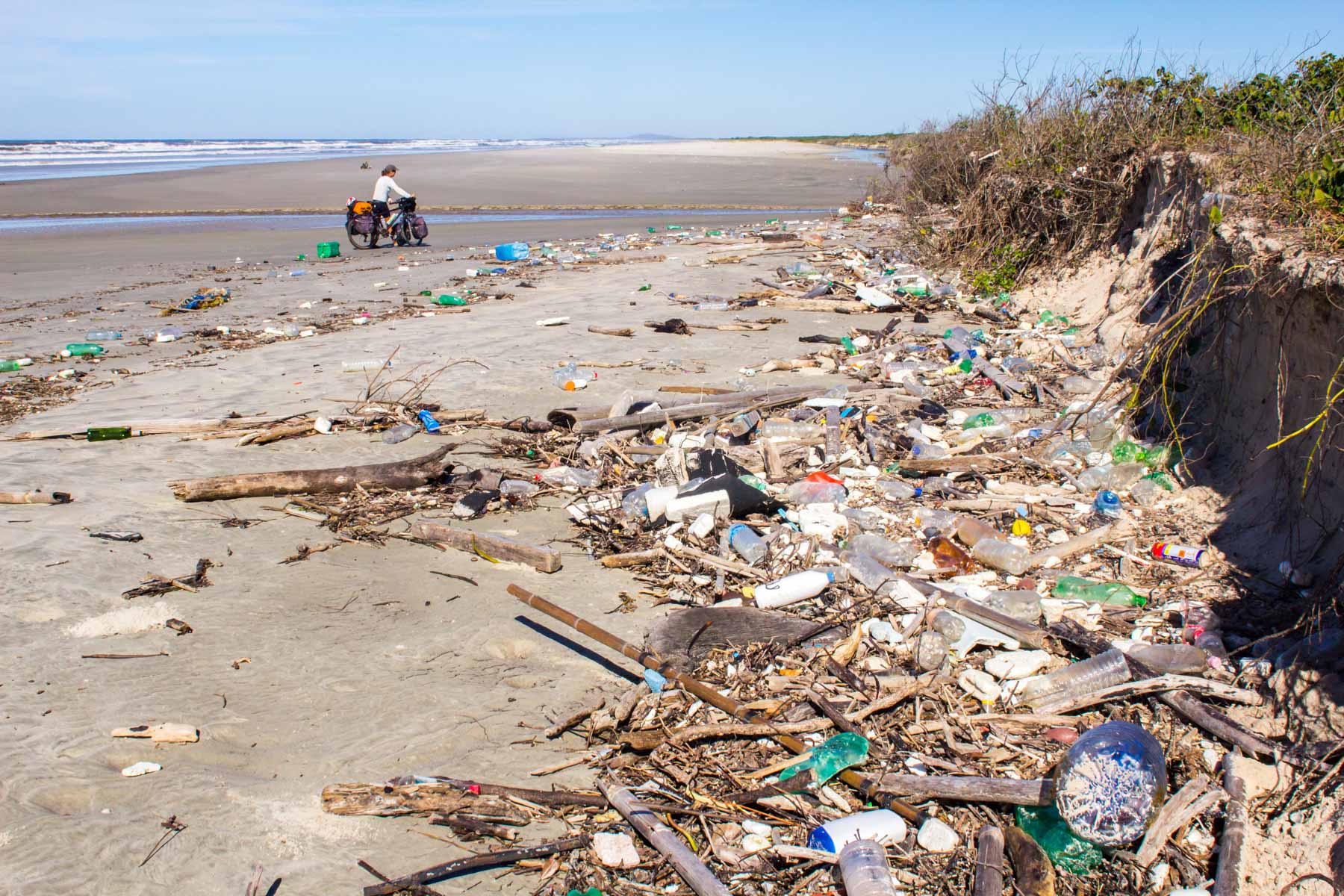 De nombreux déchets en plastique jonchent le sable d’une plage du Brésil. Au loin, on peut apercevoir, une femme avec son vélo de cyclotourisme.