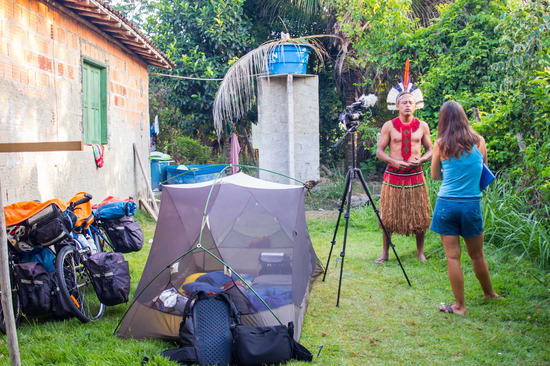 Un homme autochtone en habit traditionnel donne une entrevue filmée avec une femme de dos près d’une petite tente et d’une maison en brique.