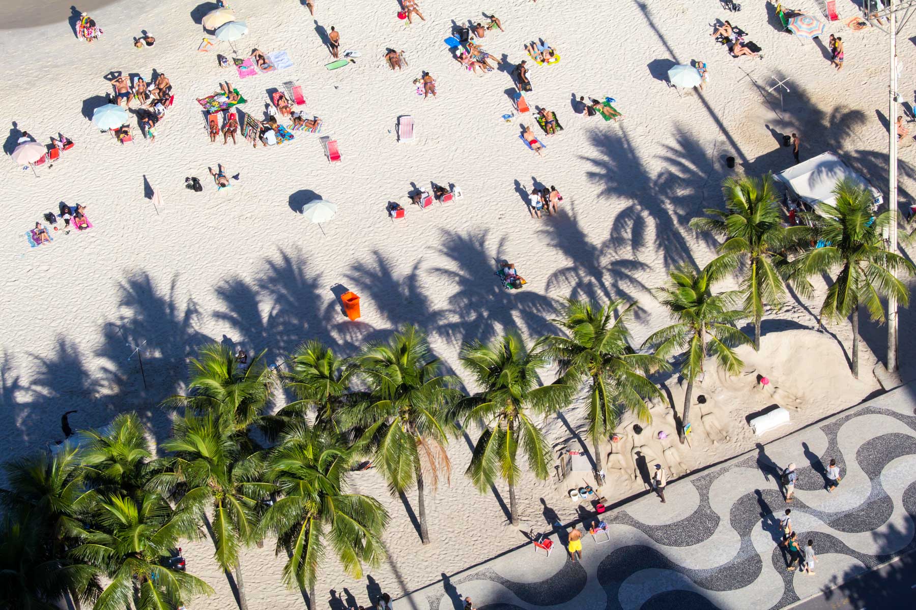 De nombreuses personnes se prélassent sur le sable chaud près de l’ombre des palmiers de cette promenade aux pavés onduleux.