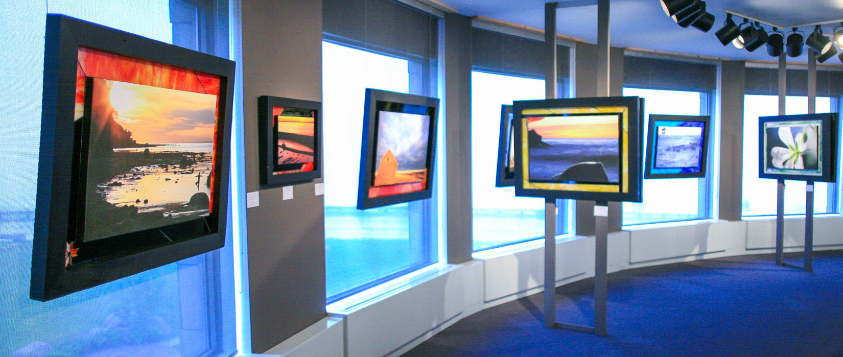 Oeuvres artistiques exposées dans une salle d’exposition avec de grandes fenêtres. Au premier plan, une œuvre représentant la silhouette d’une femme en équilibre au bord de l’eau au lever du soleil.