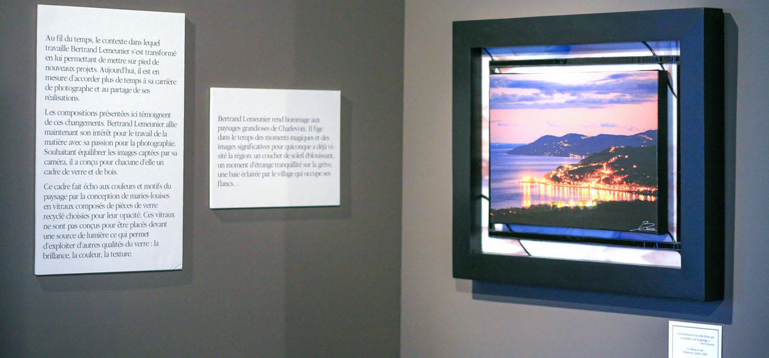 Détails d’une salle d’exposition avec des textes au mur et une photo imprimée sur une toile de peinture entourée de vitraux colorés.