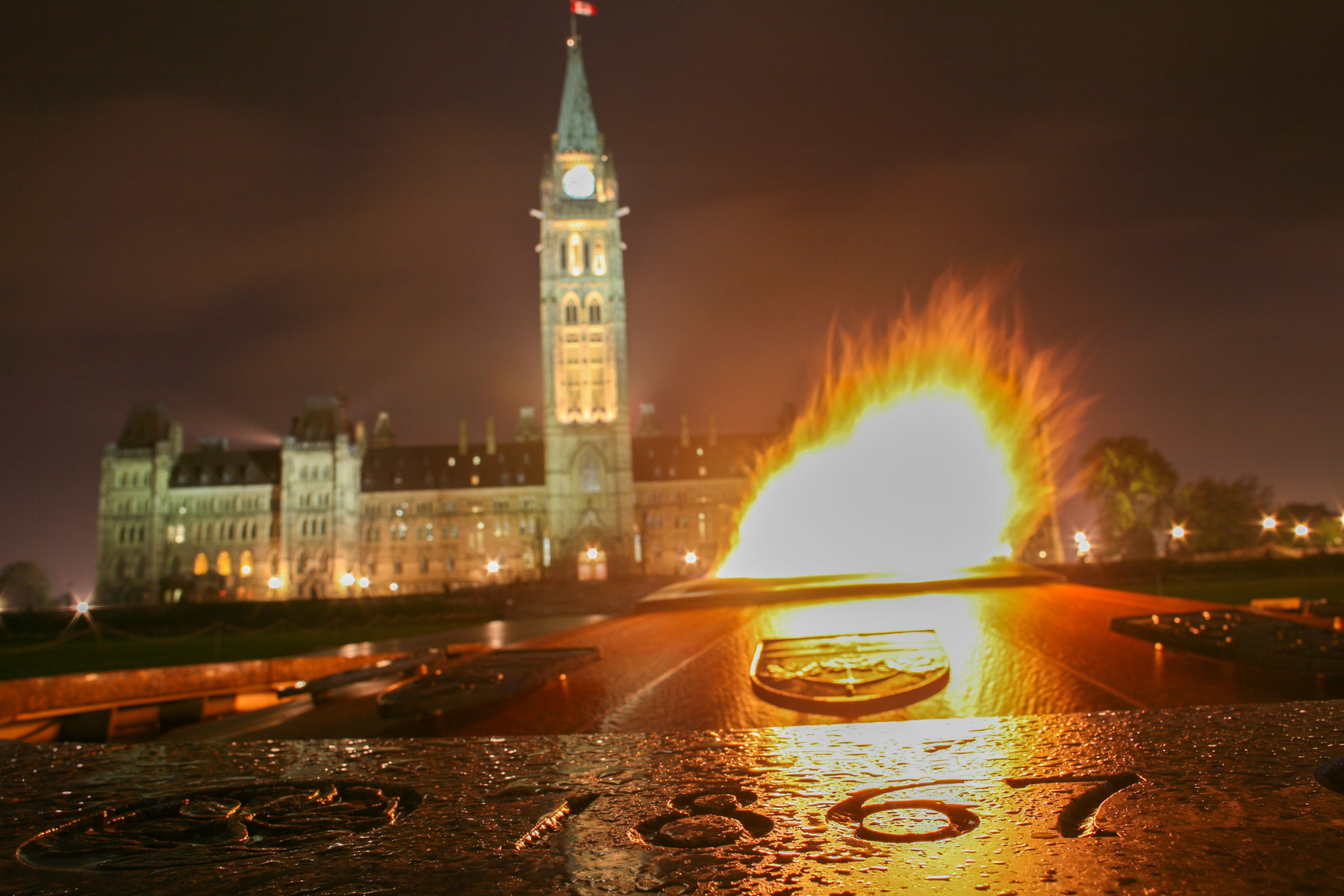 De nuit, une flamme brûle au centre d’une fontaine circulaire avec en arrière plan, un imposant bâtiment néogothique avec une haute tour.