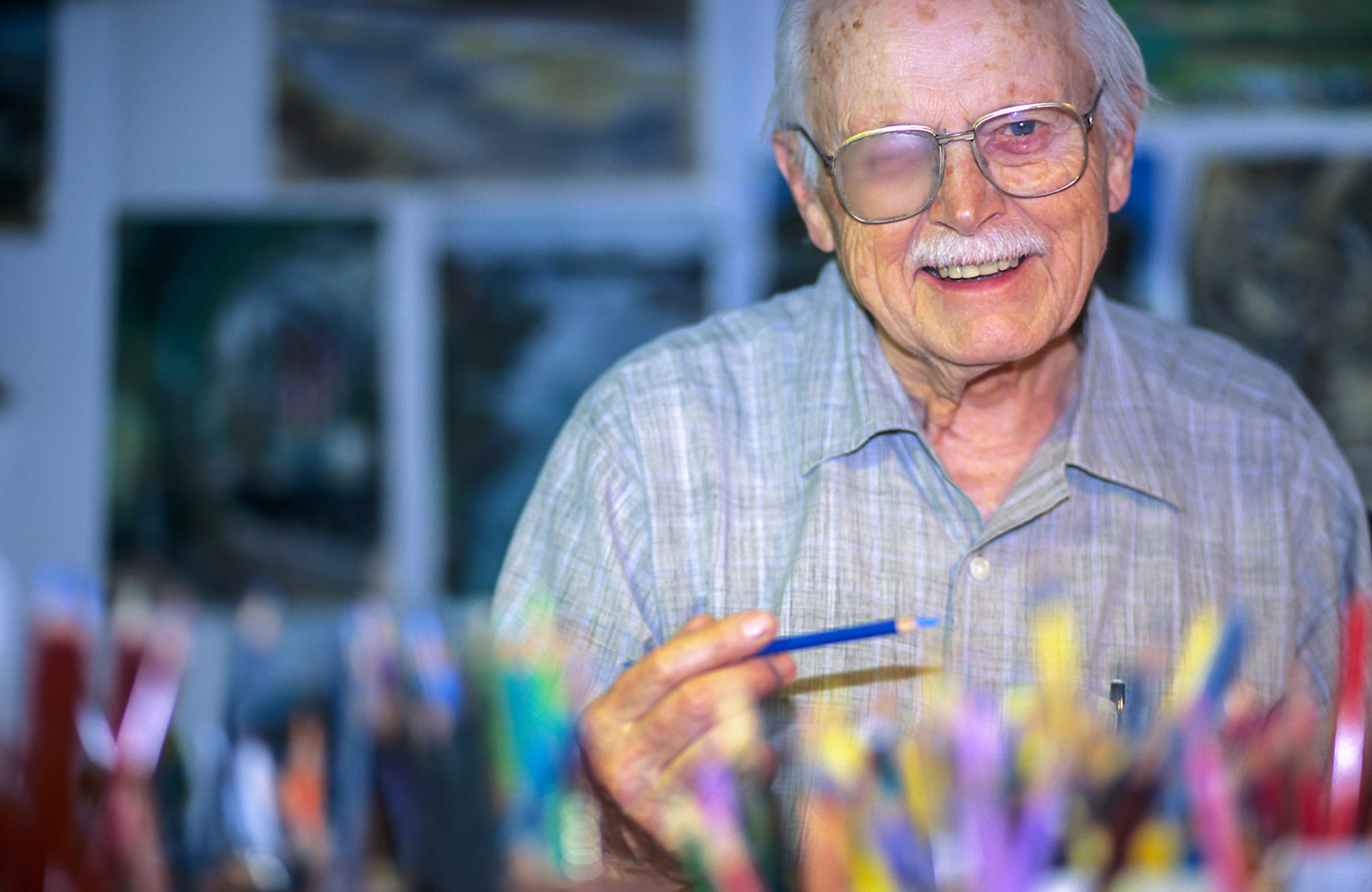 Portrait d’un artiste aux cheveux blanc et à la moustache blanche posant près de ses crayons de couleur.