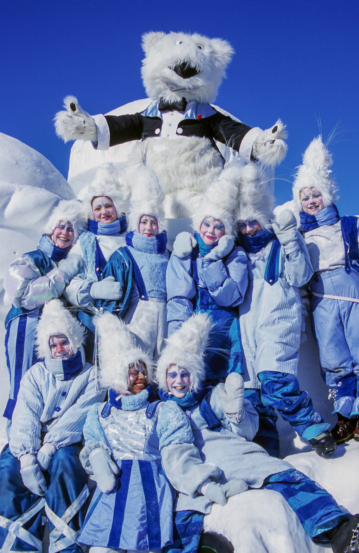 Une mascotte d’ours polaire pose avec une dizaine de jeunes femmes déguisées avec des costumes bleus et de hauts bonnets blancs.