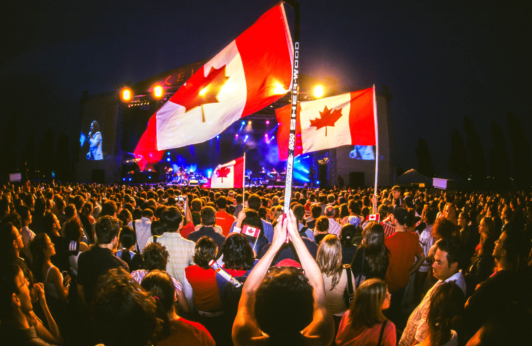 Trois grands drapeaux blanc et rouge se dressent au-dessus d’une foule amassée près d’une scène de spectacle éclairée la nuit.