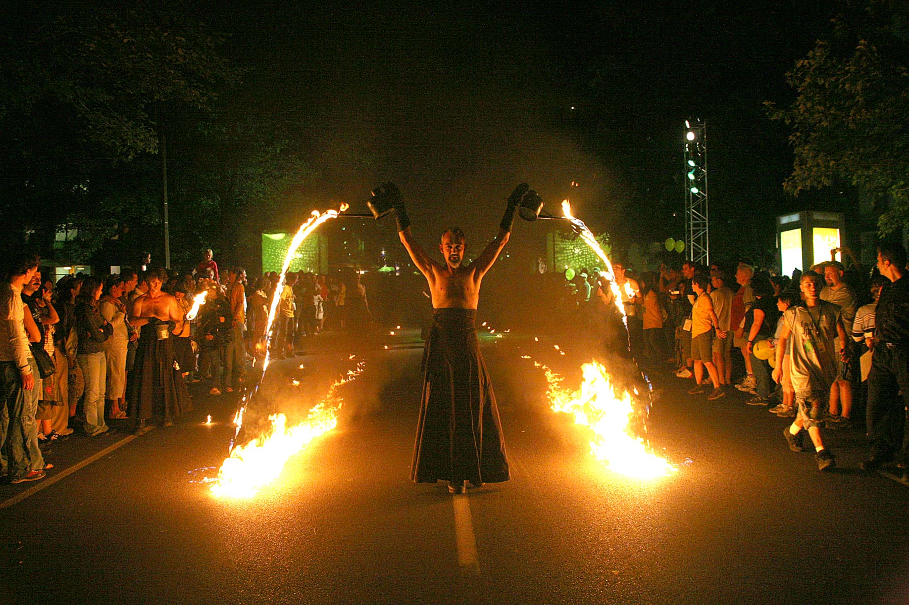 Un homme marche de nuit au milieu de la rue en tenant dans les airs deux arrosoirs d’où s’écoule un produit inflammable en feu.