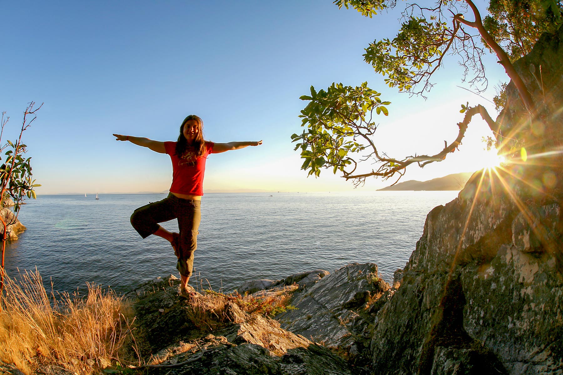 Sur des roches au bord de l’océan Pacifique, une femme se tient en équilibre sur une jambe au coucher du soleil.