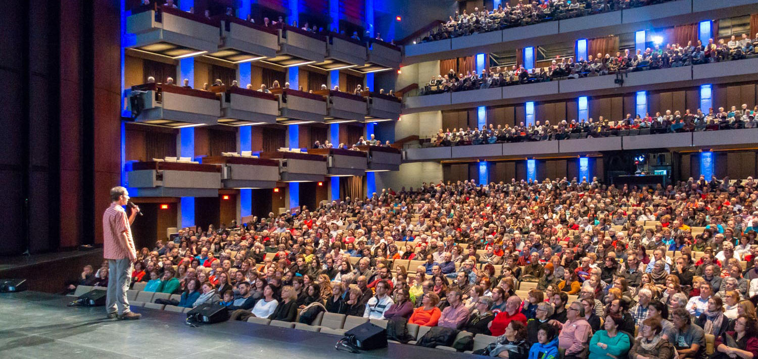 Dans un grand théâtre, un homme sur scène parle au micro devant une foule de près de 2000 personnes.