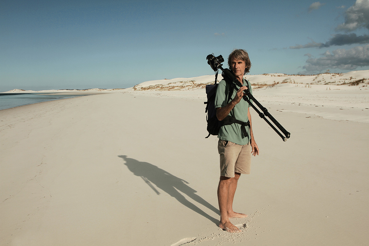 Sur une plage de sable, un grand photographe mince porte un trépied photo avec sa caméra sur son épaule droite.