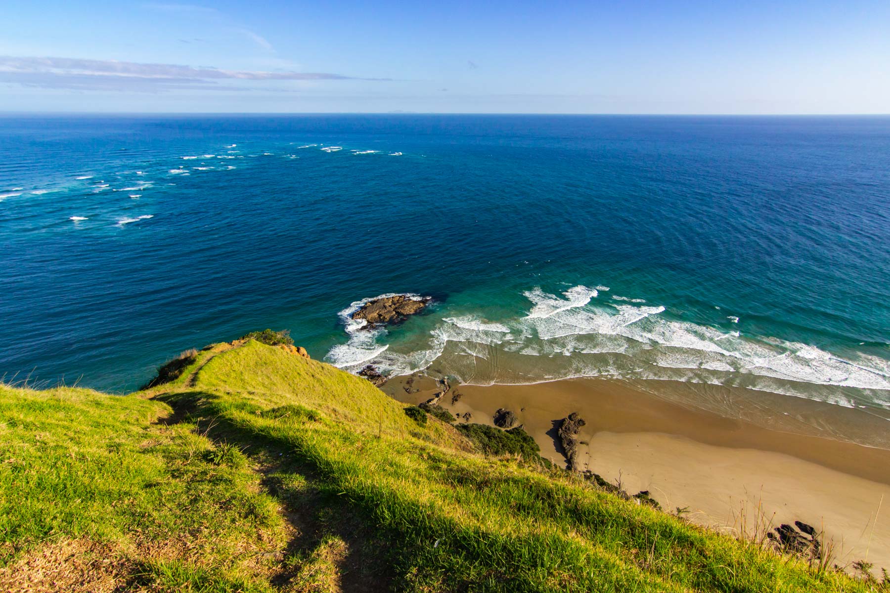 La pointe du Cap Reinga, une colline verdoyante, descend vers une petite plage de sable. Devant nous, la mer de Tasman à gauche rejoint l’océan Pacifique à droite.