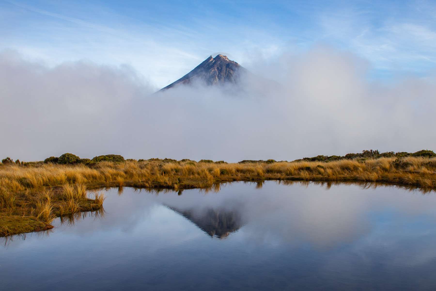 Le sommet d’un volcan dépasse les nuages et se reflète sur les eaux paisibles d’un petit étang bordé par de hautes herbes jaunes.