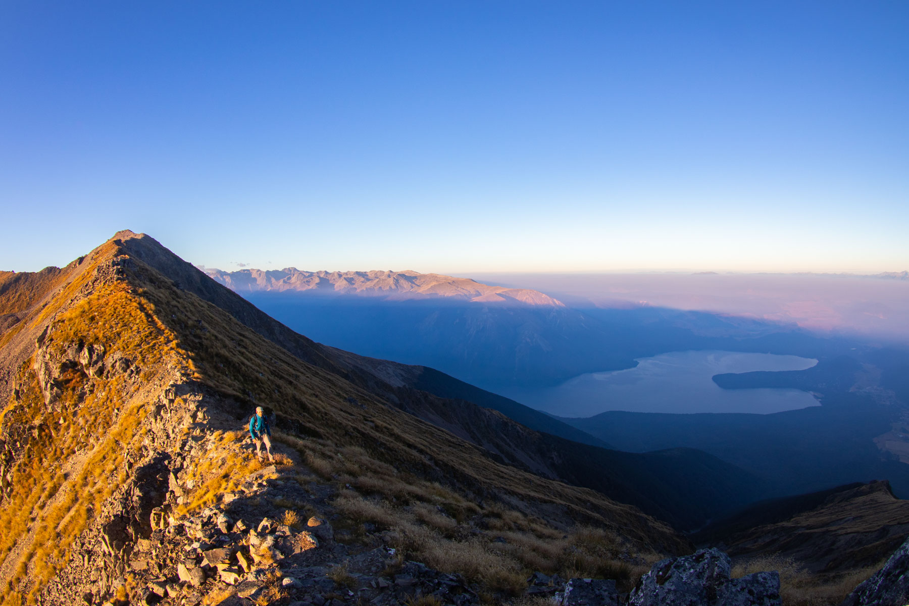 Au lever du soleil, un randonneur marche sur la crête d’une montagne. Les contours d’un lac se dessinent dans le bas de la vallée.