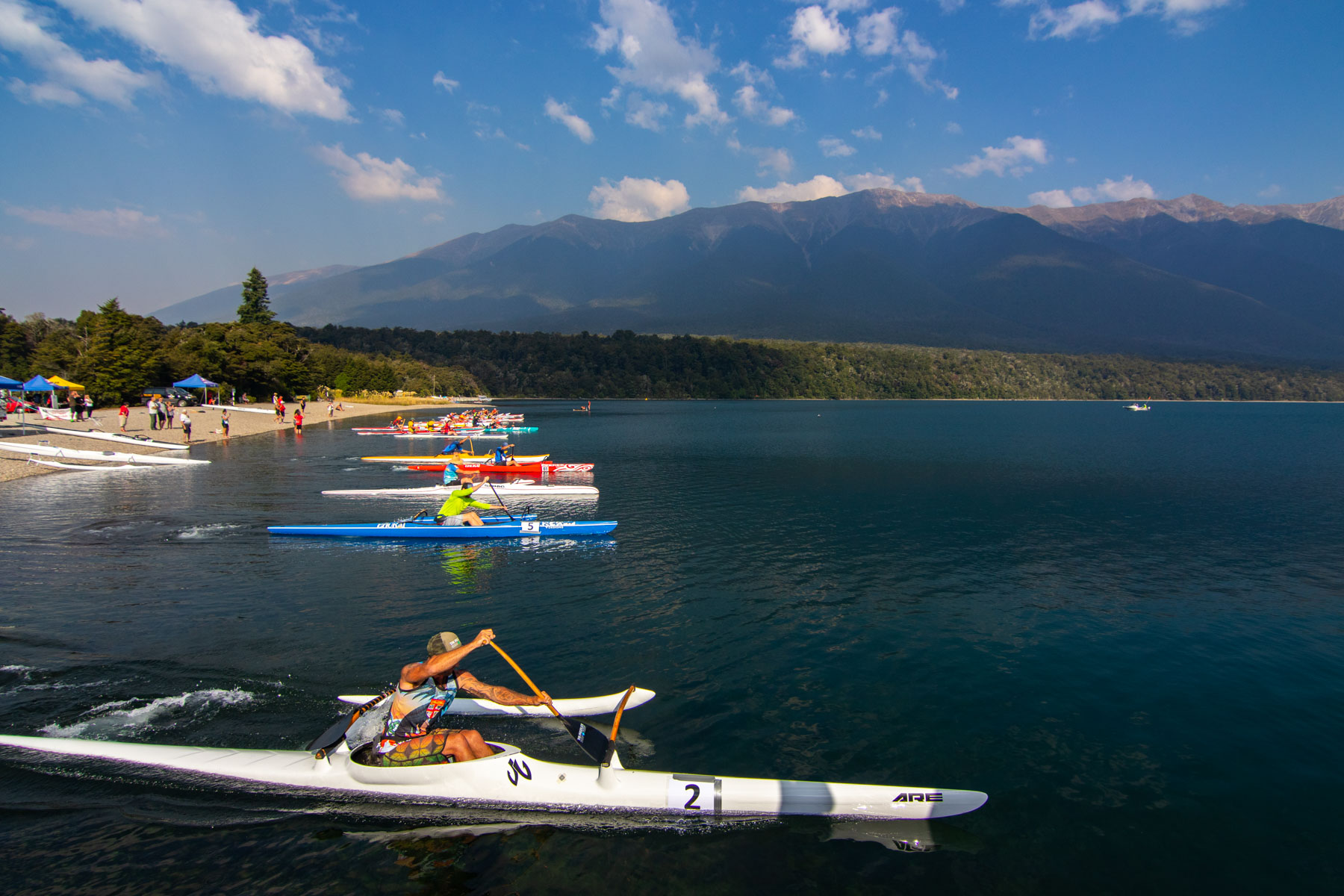 Une dizaine de canots modernes et colorés débutent une course depuis le rivage d’un lac aux pieds des montagnes.