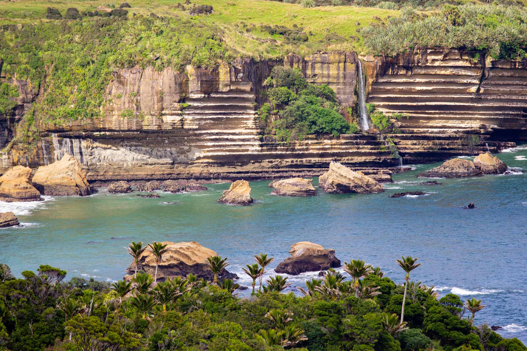 Une cascade coule le long d’une falaise aux strates horizontales tandis que des vagues se brisent sur des îlots rocheux.