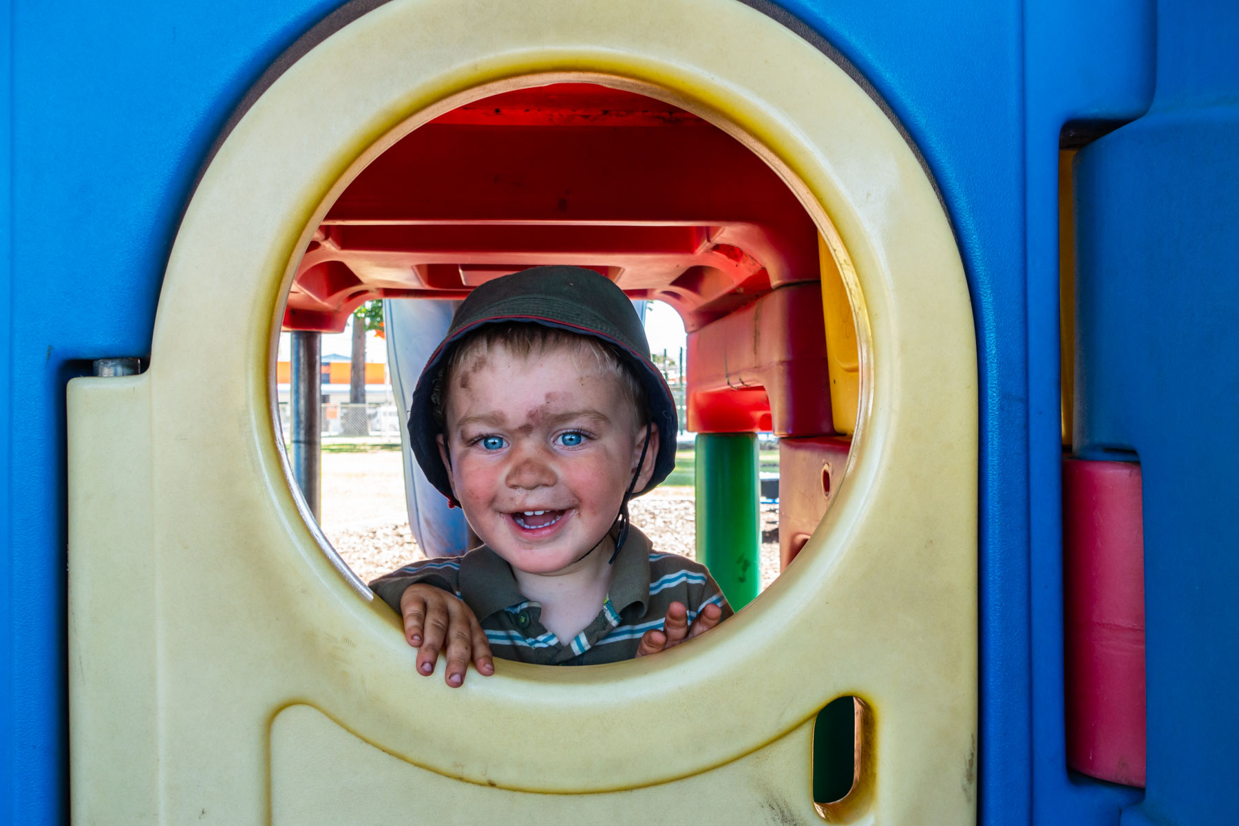 Le visage barbouillé de terre, un jeune garçon sourit au photographe par l'ouverture d'un module de jeux jaune et bleu.