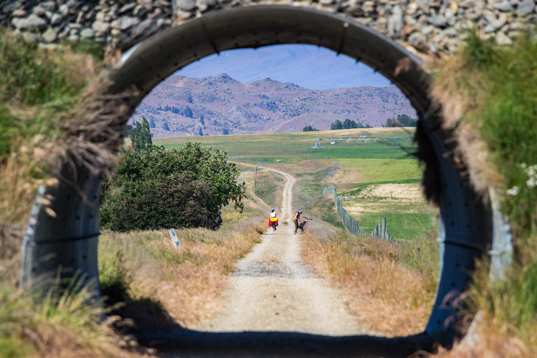 Paysage de campagne où un sentier passe à travers un petit tunnel puis continue en ligne droite. Au loin, on distingue deux cyclistes, des arbres et des collines rocailleuses.