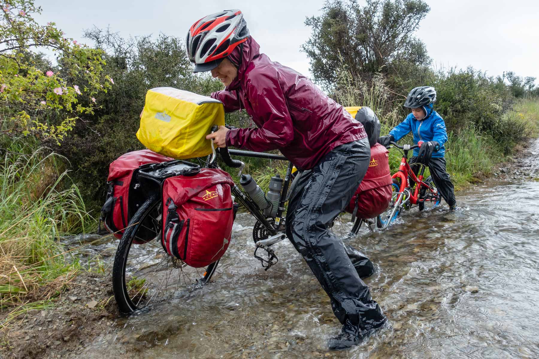Une femme traverse un ruisseau en poussant son vélo chargé.