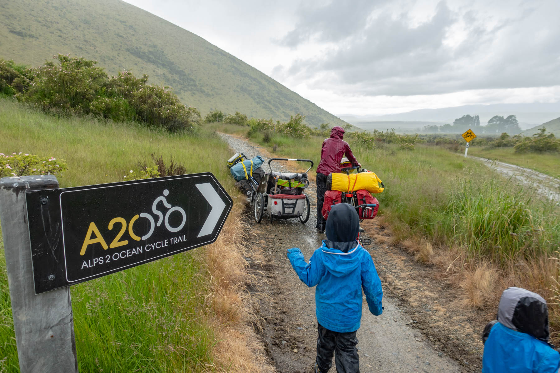 Dans un décors de montagne, deux enfants suivent à pied leur mère qui pousse son vélo sous la pluie.