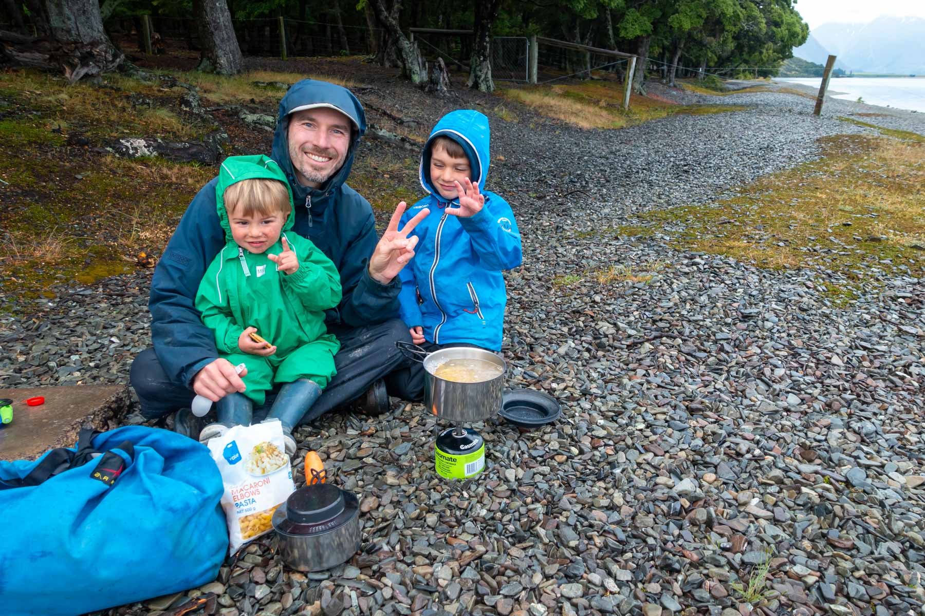 Un homme et deux enfants cuisinent avec un petit réchaud sur une plage de galets.