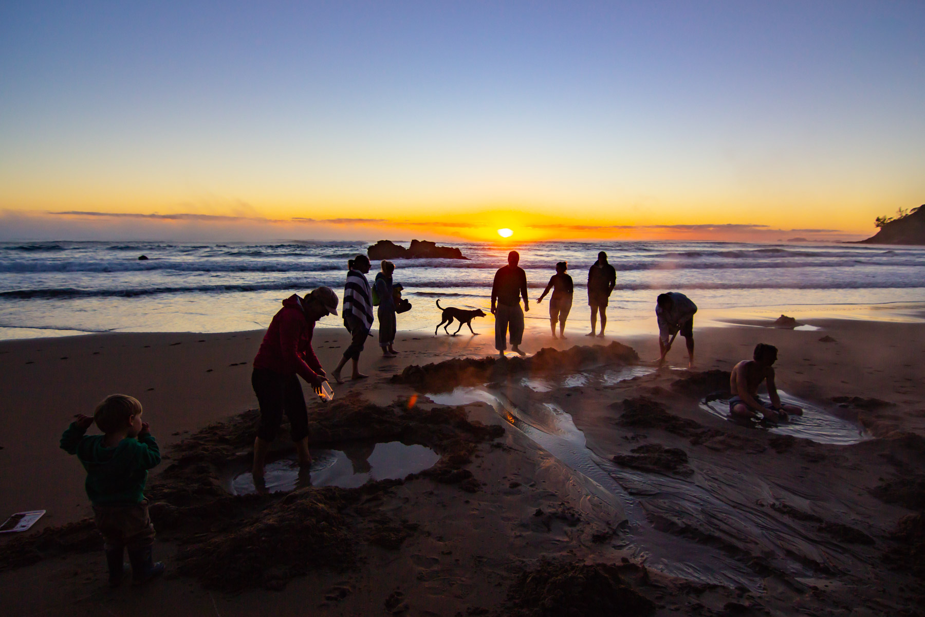 Au lever de soleil, silhouettes de personnes debout sur une plage. Certaines se creusent dans le sable une petite piscine d’eau chaude.