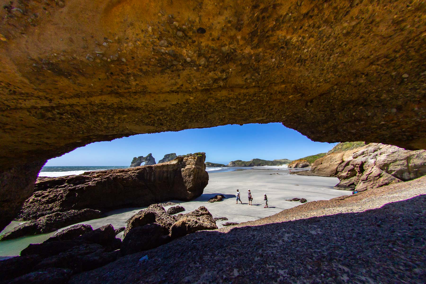 Une imposante arche rocheuse se dresse au premier plan tandis que plusieurs silhouettes arpentent une plage de sable parsemé d’îlots rocheux.