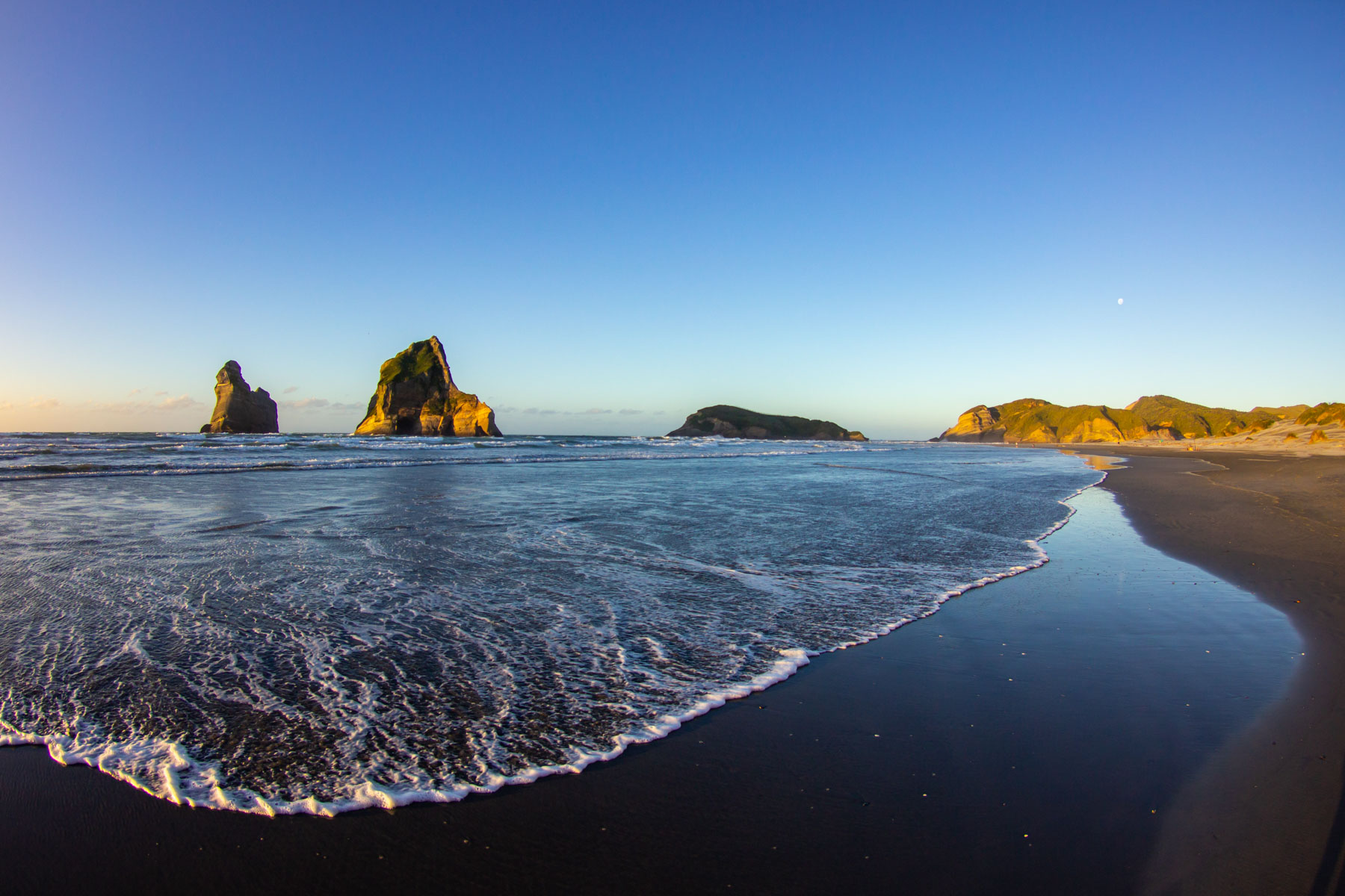 Sous un ciel bleu, une vague glisse en douceur sur une longue plage de sable. Plusieurs îlots rocheux apparaissent sur la ligne d’horizon.