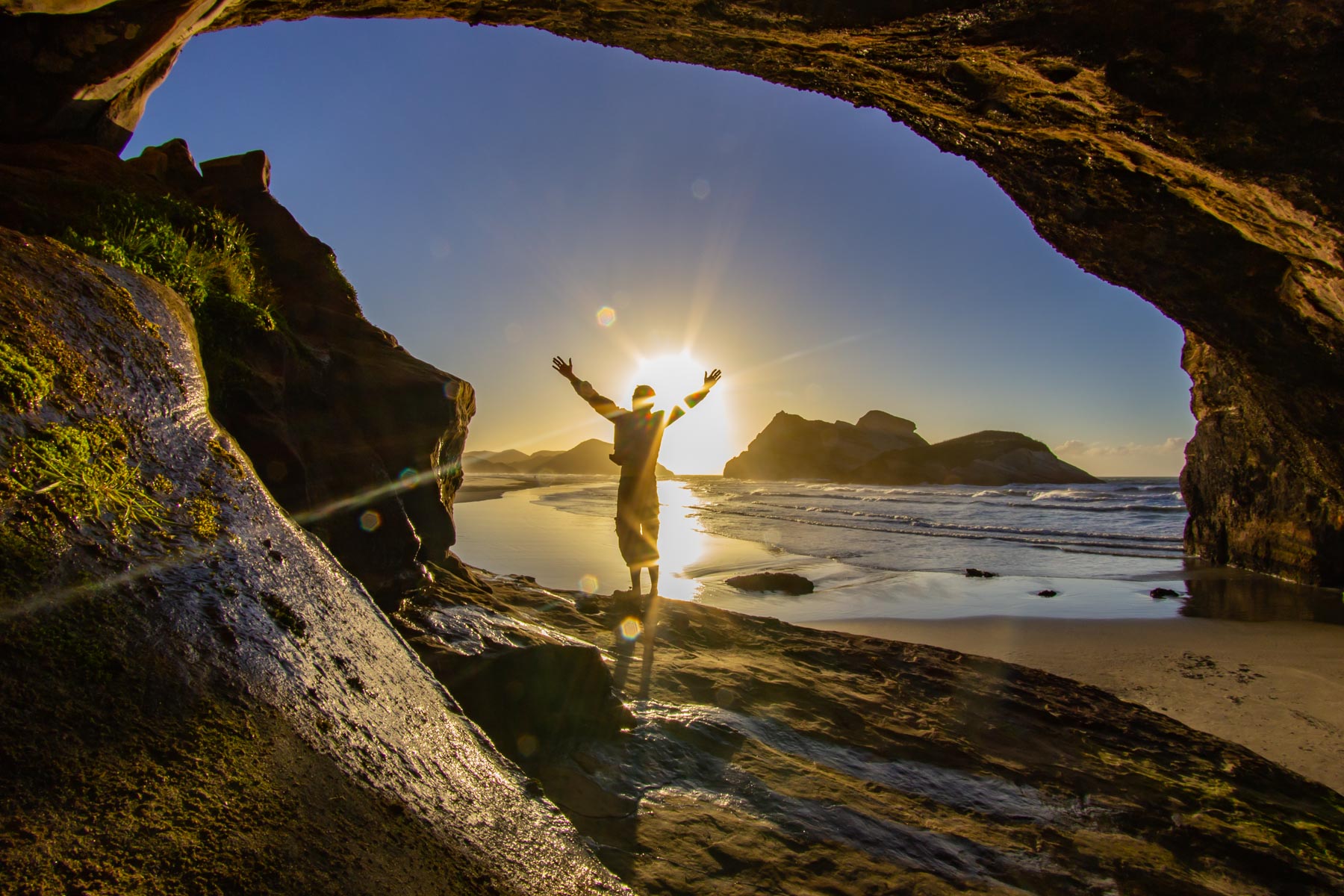 Silhouette d’une personne les bras en l’air sous une arche rocheuse près d’une plage de sable au coucher du soleil.
