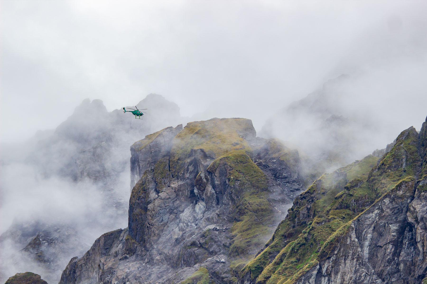 Un hélicoptère vert s’envole vers les sommets nuageux.