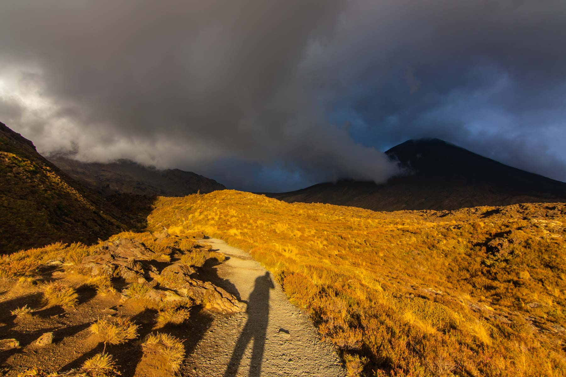 L’ombre du photographe se dessine sur le sol au coucher du soleil alors que des nuages noirs engloutissent le volcan Ngauruhoe.