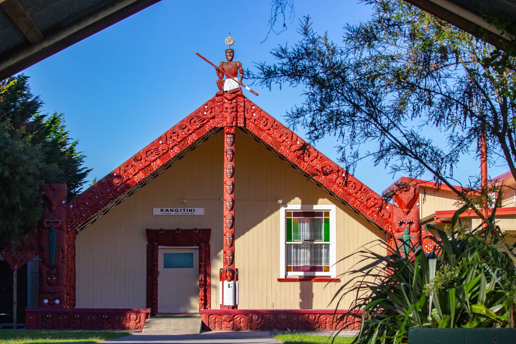 Façade d’un bâtiment de forme triangulaire dont la structure présente des sculptures traditionnelles māories peintes en rouge.