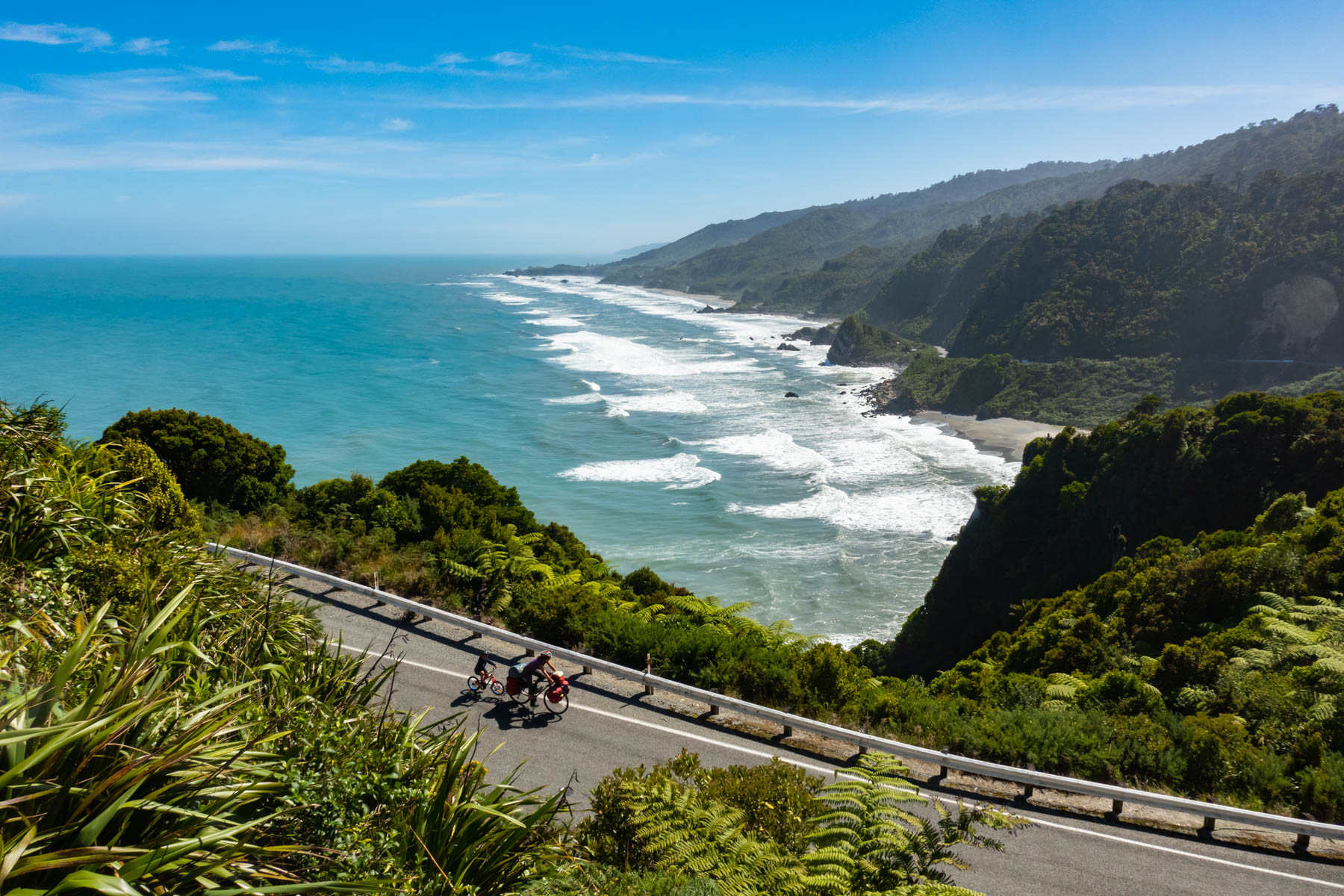 Une femme et son fils à vélos empruntent une route longeant la côte Ouest de l’île du sud en Nouvelle-Zélande.