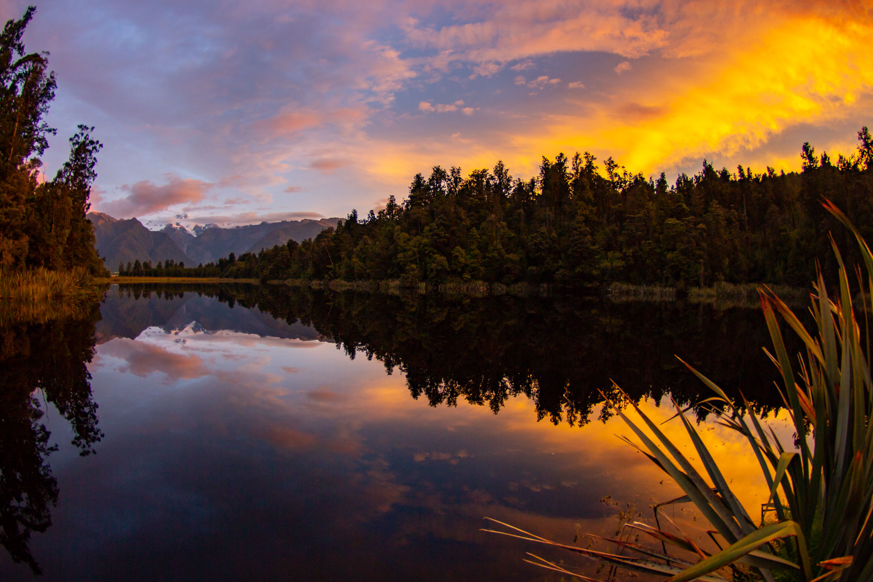 Sunset, with orange clouds reflecting on Lake Matheson and snow-capped mountains in the distance.