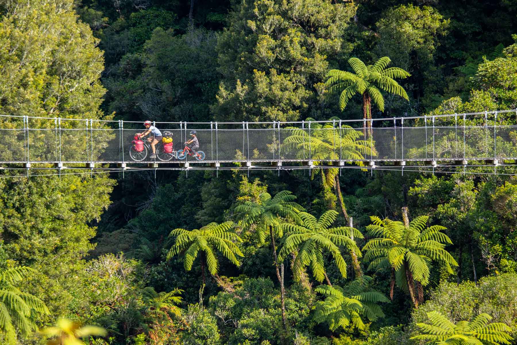 Une femme et son fils roulent à vélo sur un pont suspendu entouré d’une végétation luxuriante.